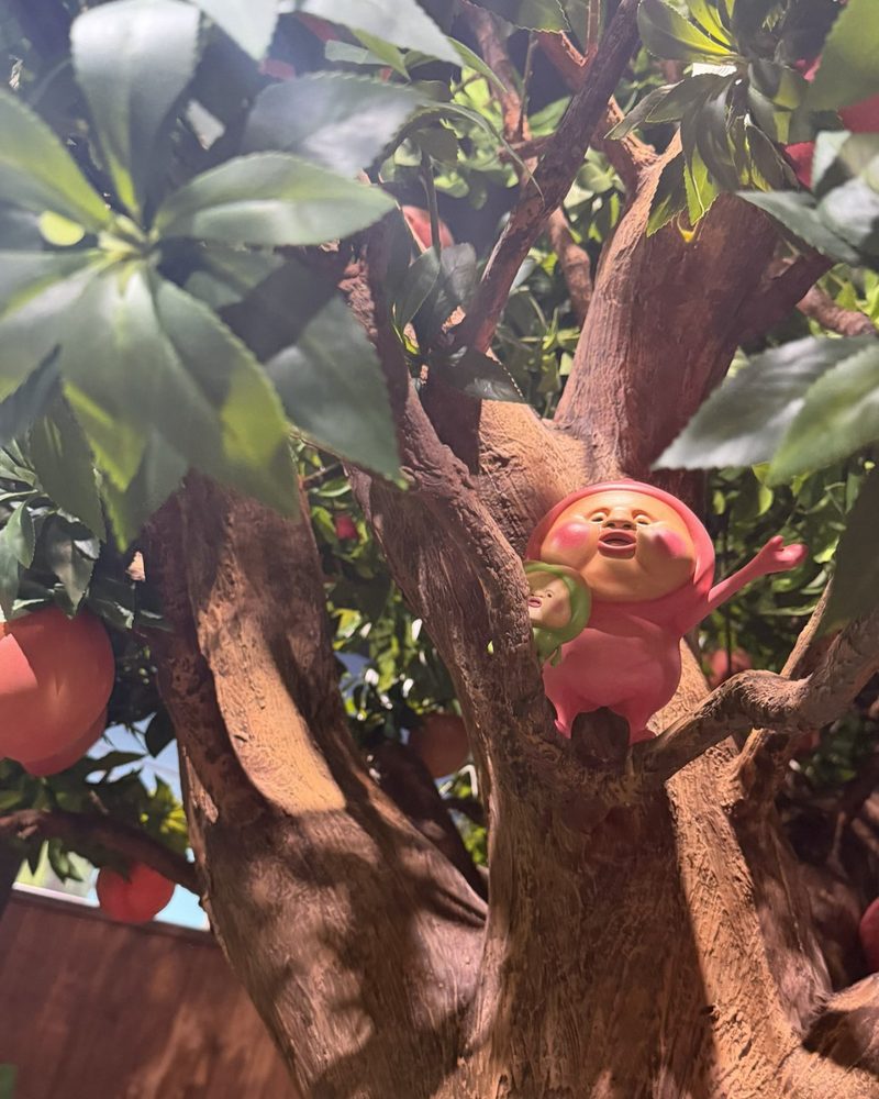 A pink Kobito figure with a round face sitting in the branches of a peach tree