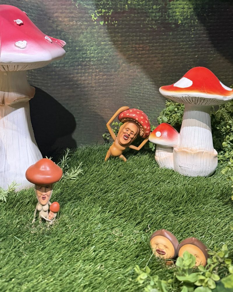 Kobito figures with mushroom caps standing on artificial grass among big mushrooms
